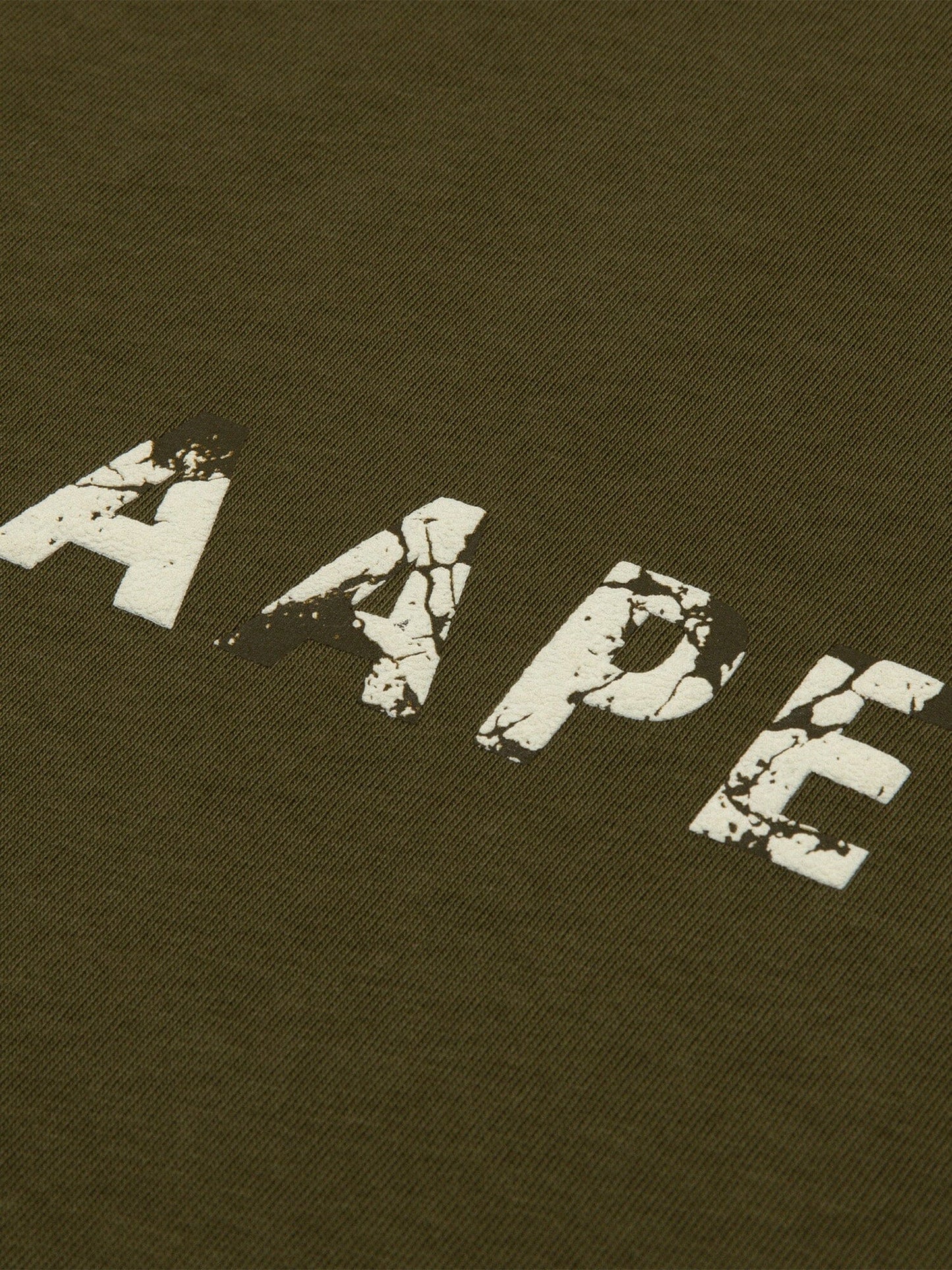 Close-up of the AAPE X Alpha Somewhere In Universe Tee by Alpha Industries. Olive green fabric with AAPE in distressed white font. 