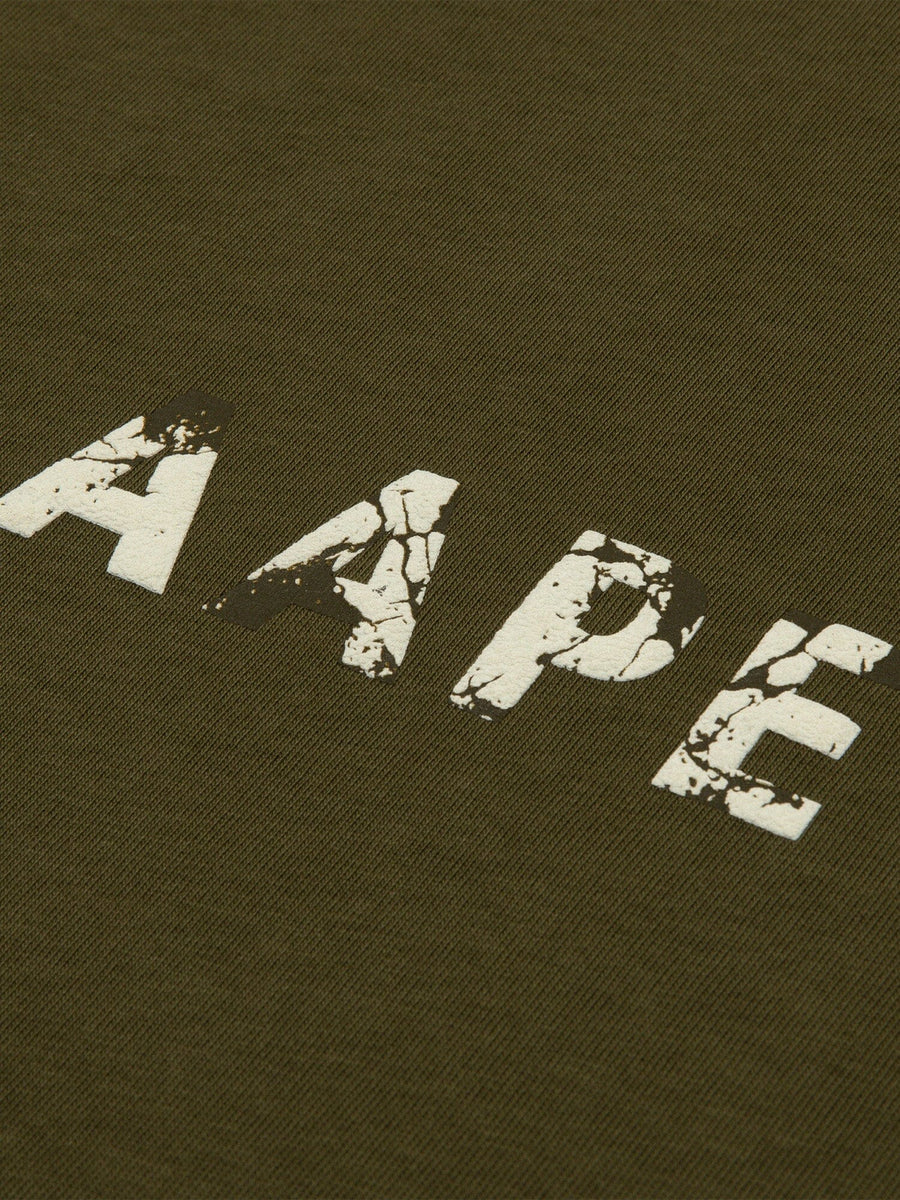Close-up of the AAPE X Alpha Somewhere In Universe Tee by Alpha Industries. Olive green fabric with AAPE in distressed white font. 