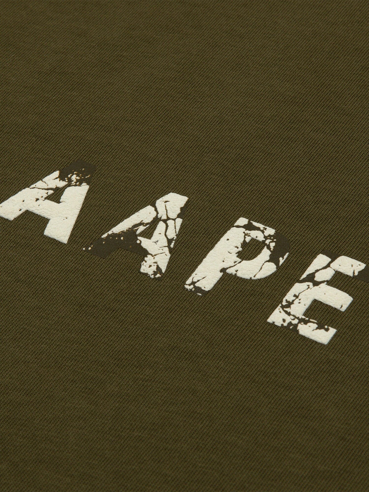 Close-up of the AAPE X Alpha Somewhere In Universe Tee by Alpha Industries. Olive green fabric with AAPE in distressed white font. 