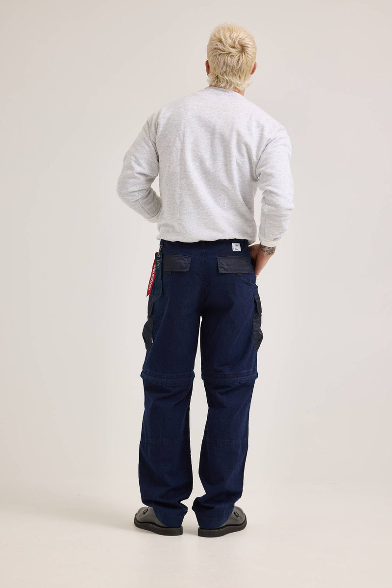 A person with a blond mullet is wearing a gray long-sleeve shirt, black shoes, and dark blue Alpha X Lee Convertible Cargo Pant from Alpha Industries, facing away.