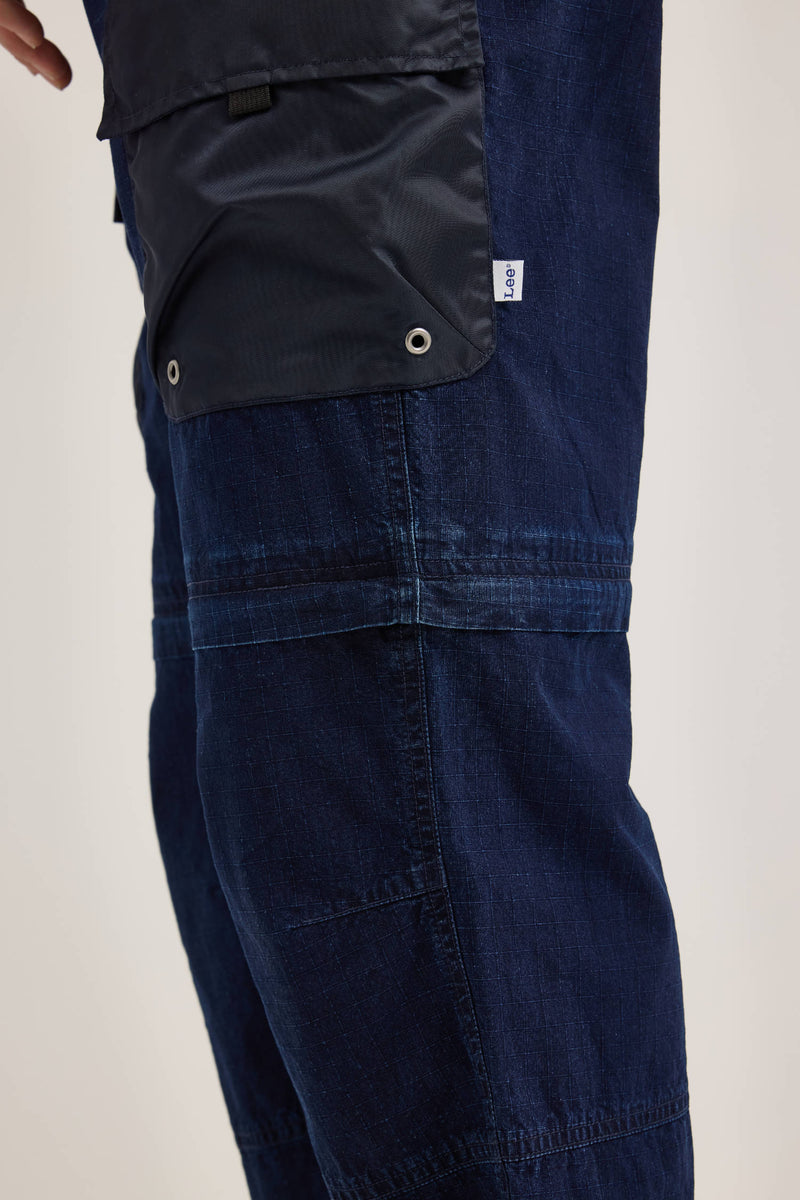 Close-up of a person wearing the Alpha X Lee Convertible Cargo Pant by Alpha Industries in dark blue denim, showcasing a removable black pocket flap fastened with silver snaps, epitomizing the convertible cargo pants design.