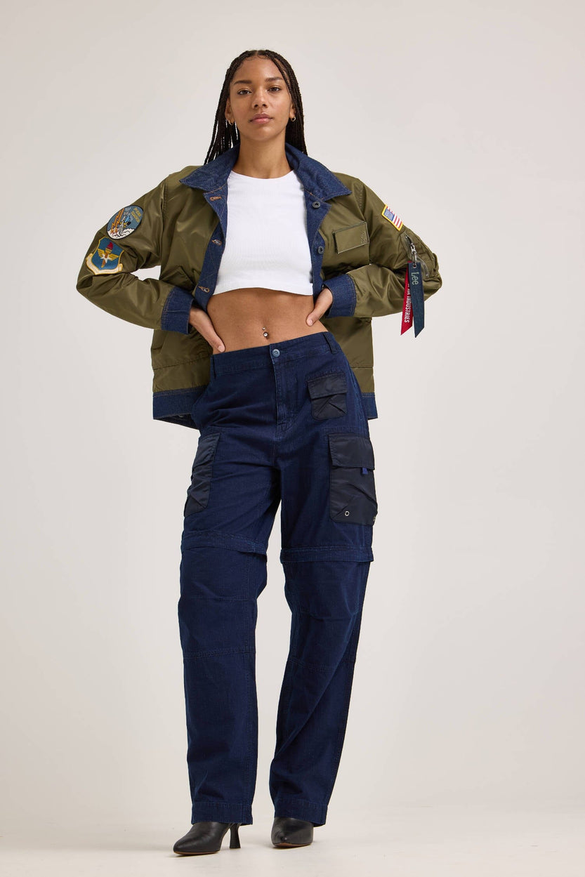 A person wearing a green jacket and white crop top stands confidently, sporting Alpha Industries x FW24-Lee's Alpha x Lee Convertible Cargo Pant in blue ripstop cotton, completing the look with black shoes.