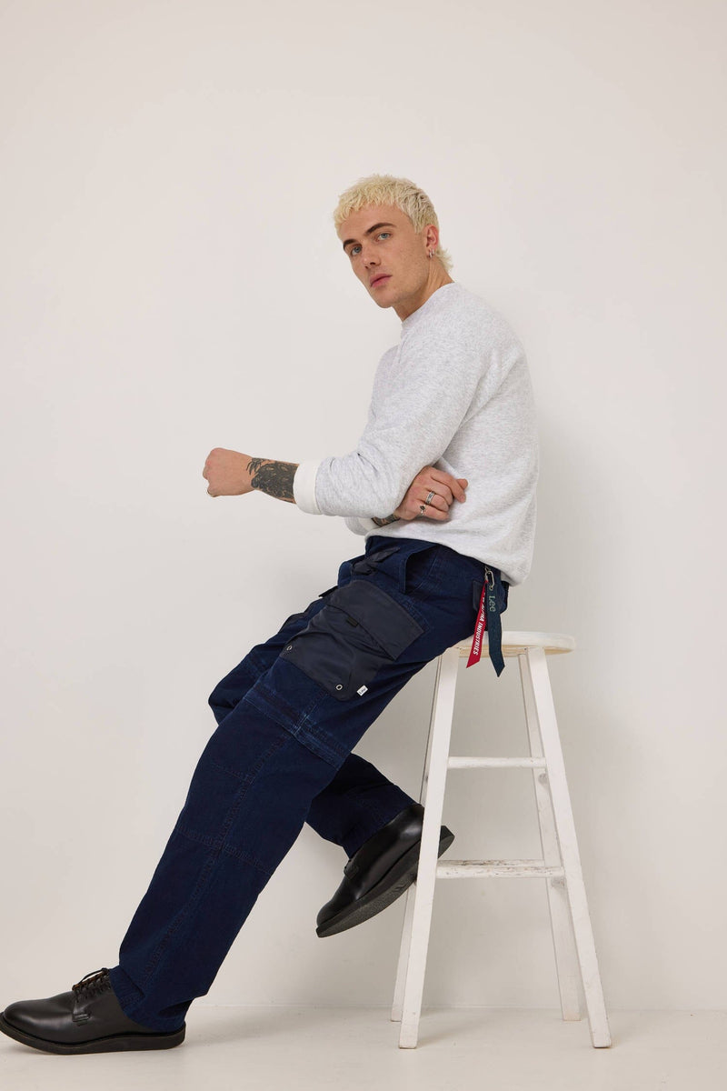 A person with short blond hair sits on a white stool, wearing a white long-sleeve shirt and the stylish Alpha X Lee Convertible Cargo Pant from Alpha Industries, crafted from ripstop cotton. They're sporting black shoes, effortlessly showcasing the Alpha x Lee collaboration.