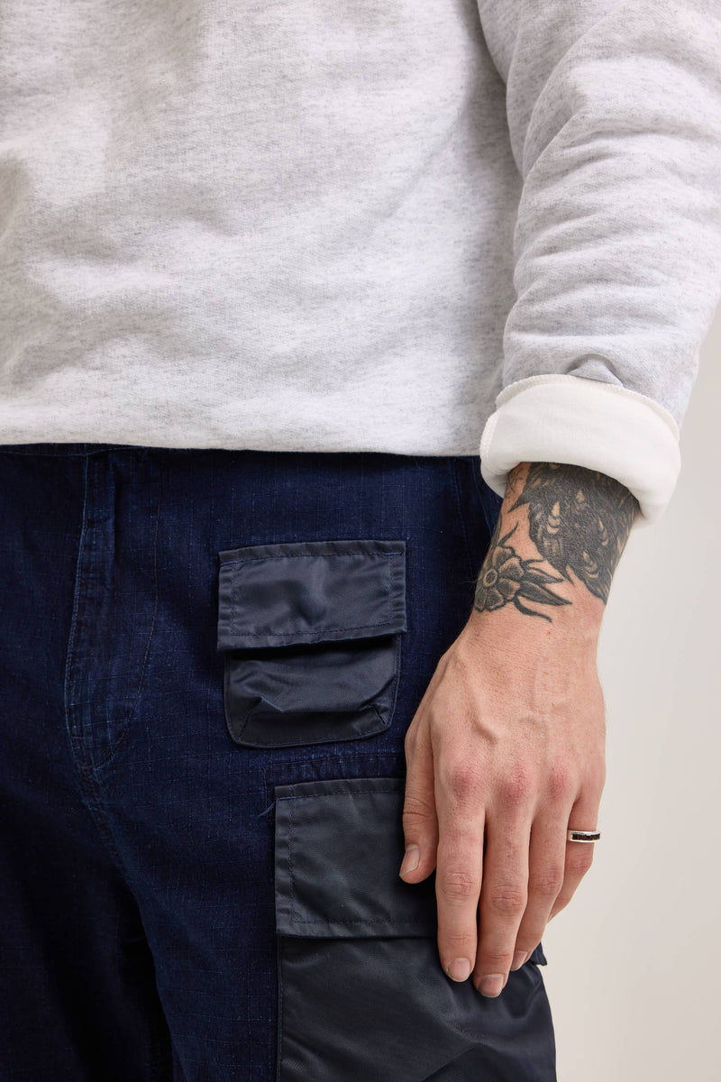 A person is wearing a gray sweatshirt with rolled-up sleeves and dark Alpha X Lee Convertible Cargo Pants, featuring a tattoo and an Alpha Industries/Lee ring on their left hand.