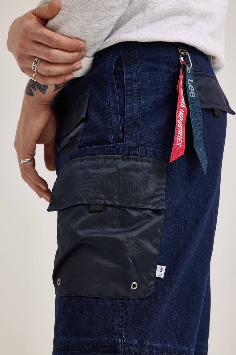 Side view of a person wearing the Alpha X Lee Convertible Cargo Pant by Alpha Industries, crafted from dark blue ripstop cotton, featuring a black pocket and red keychain. The individual sports a gray shirt, adorned with rings and a tattoo on their hand.