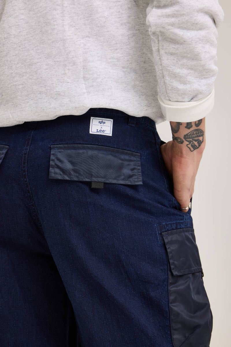 From behind, a person in a gray sweater and blue pants with cargo pockets is visible. The back of the Alpha X Lee Convertible Cargo Pant, made from durable ripstop cotton, features a label from Alpha Industries.