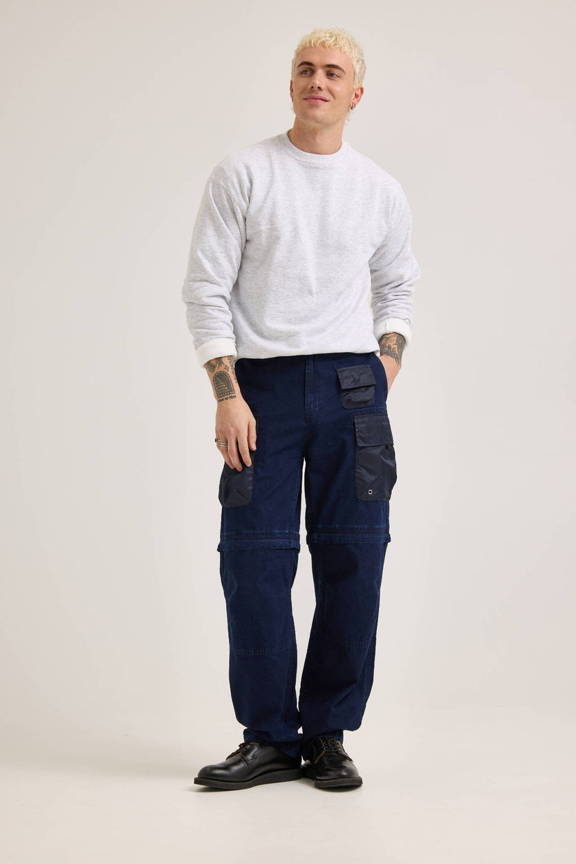 A person with short blond hair wears a gray sweatshirt and the Alpha X Lee Convertible Cargo Pant in indigo, paired with black shoes. They stand in a neutral pose.