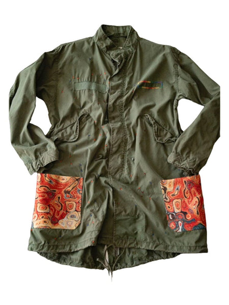 Alpha X Transnomadica Fishtail Parka by Alpha Industries, sage color with paint splatters, two large front pockets, red and orange abstract patterns.