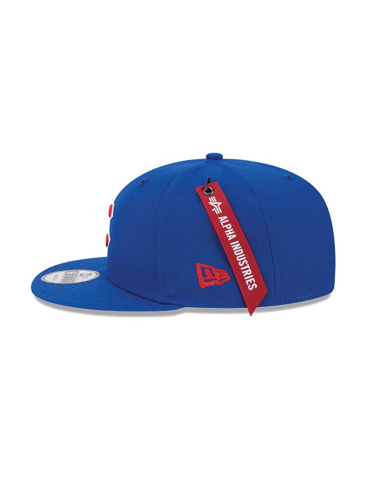 A blue Chicago Cubs cap showcasing a red embroidered logo and a trendy snapback closure. The red tag reads Alpha Industries, highlighting the collaboration between Alpha Industries and New Era®.