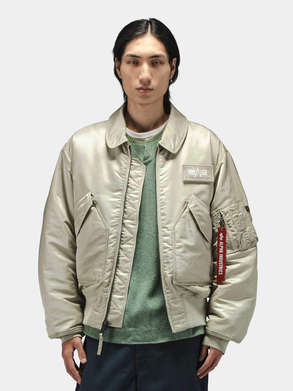 Alpha Industries Cwu 45/P Bomber Jacket in light beige, worn over a green sweater and dark pants.