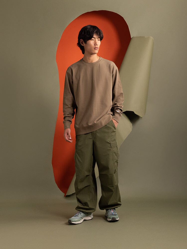 A person dressed in an Essential Crewneck in brown from Alpha Industries and olive pants is standing. The brown cotton blend unisex crewneck brings a touch of relaxed style to the scene.