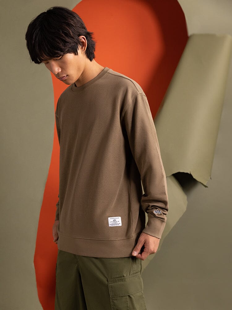 A person wearing the brown Alpha Industries Essential Crewneck and green pants, subtly adorned with the brand logo.