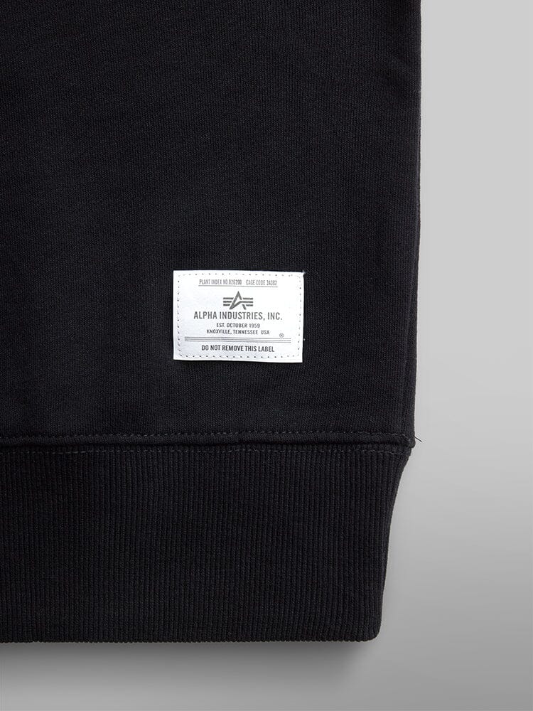 Close-Up of a black cotton blend Essential Crewneck by Alpha Industries, showcasing the label sewn at the bottom right hem.