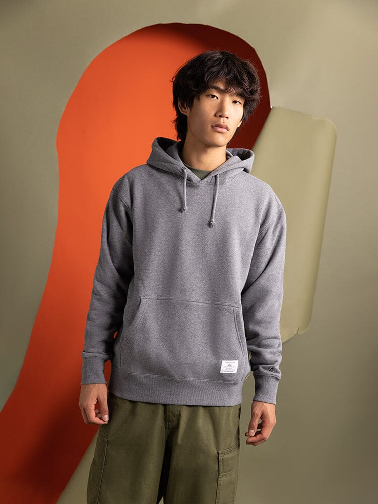 A person wearing an medium charcoal heather Essential Hoodie by Alpha Industries and green pants.
