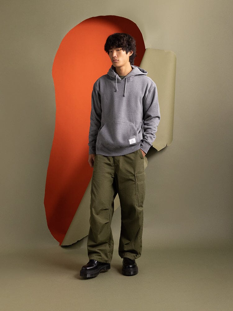 An individual is seen wearing the Essential Hoodie in medium charcoal heather from Alpha Industries, paired with green pants.