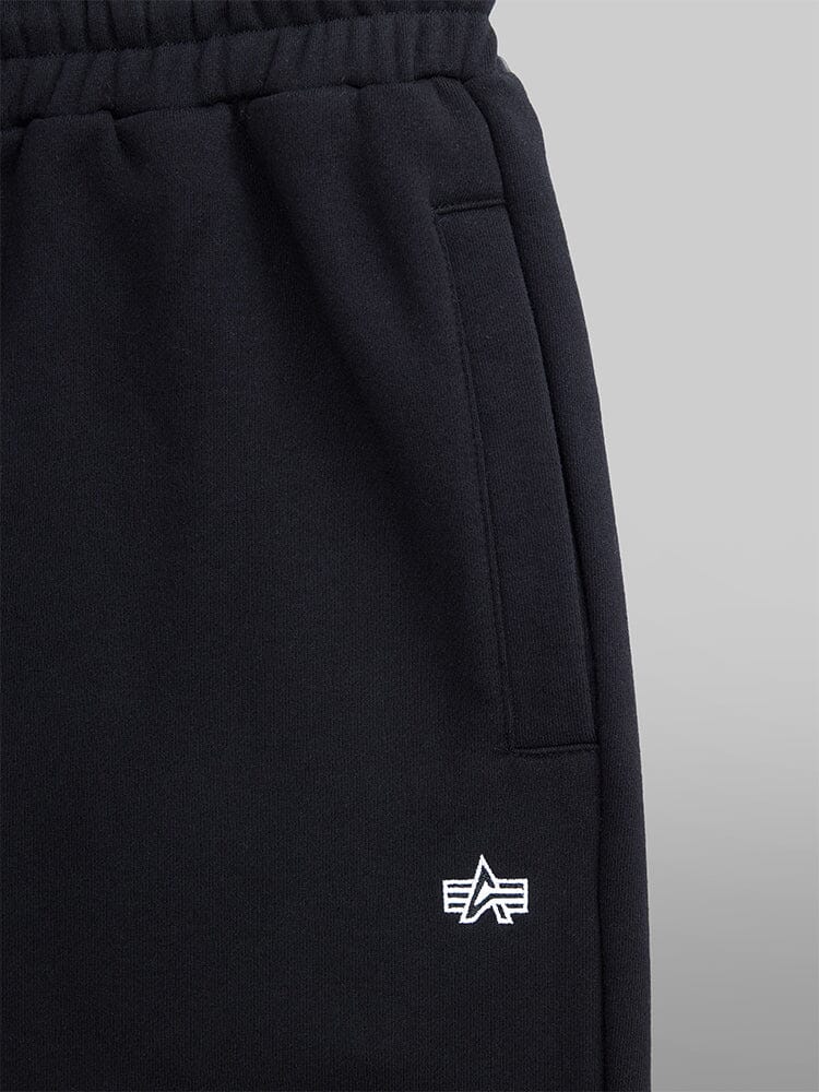 Close-up pocket view, the Women's Essential Sweatpants from Alpha Industries are designed in black with a comfortable elastic waistband. Made from soft cotton fabric, they feature a small star and stripes logo near the pocket.