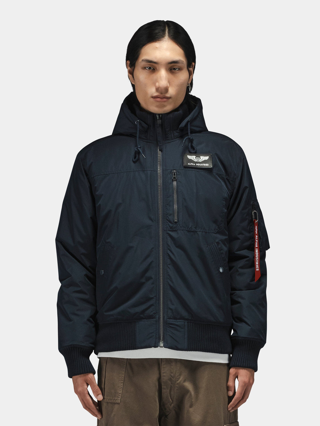 Alpha Industries Hooded Rib Flight Jacket with chest patch, zippered pocket, utility sleeve pocket with red tag, and brown pants.