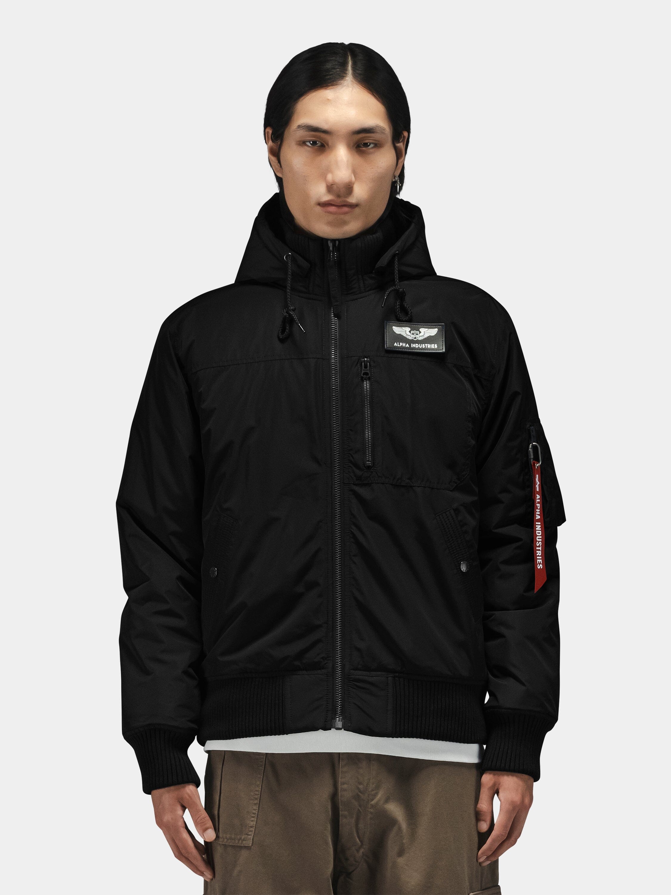 Alpha Industries Hooded Rib Flight Jacket in black with chest patch and utility sleeve pocket, paired with brown pants.
