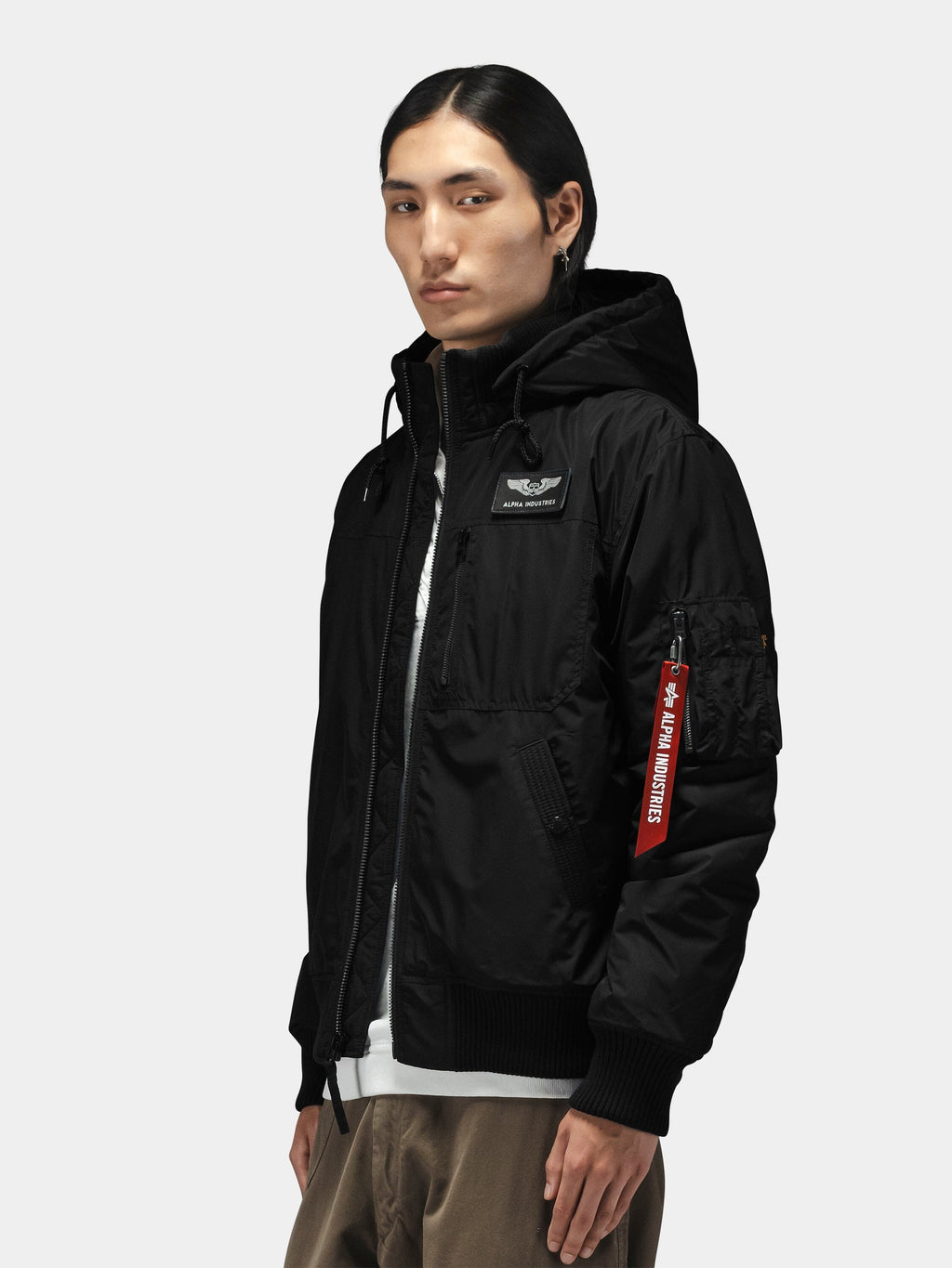 Alpha Industries Hooded Rib Flight Jacket with utility sleeve pocket, red tag, ribbed cuffs, layered over white shirt and khaki pants.