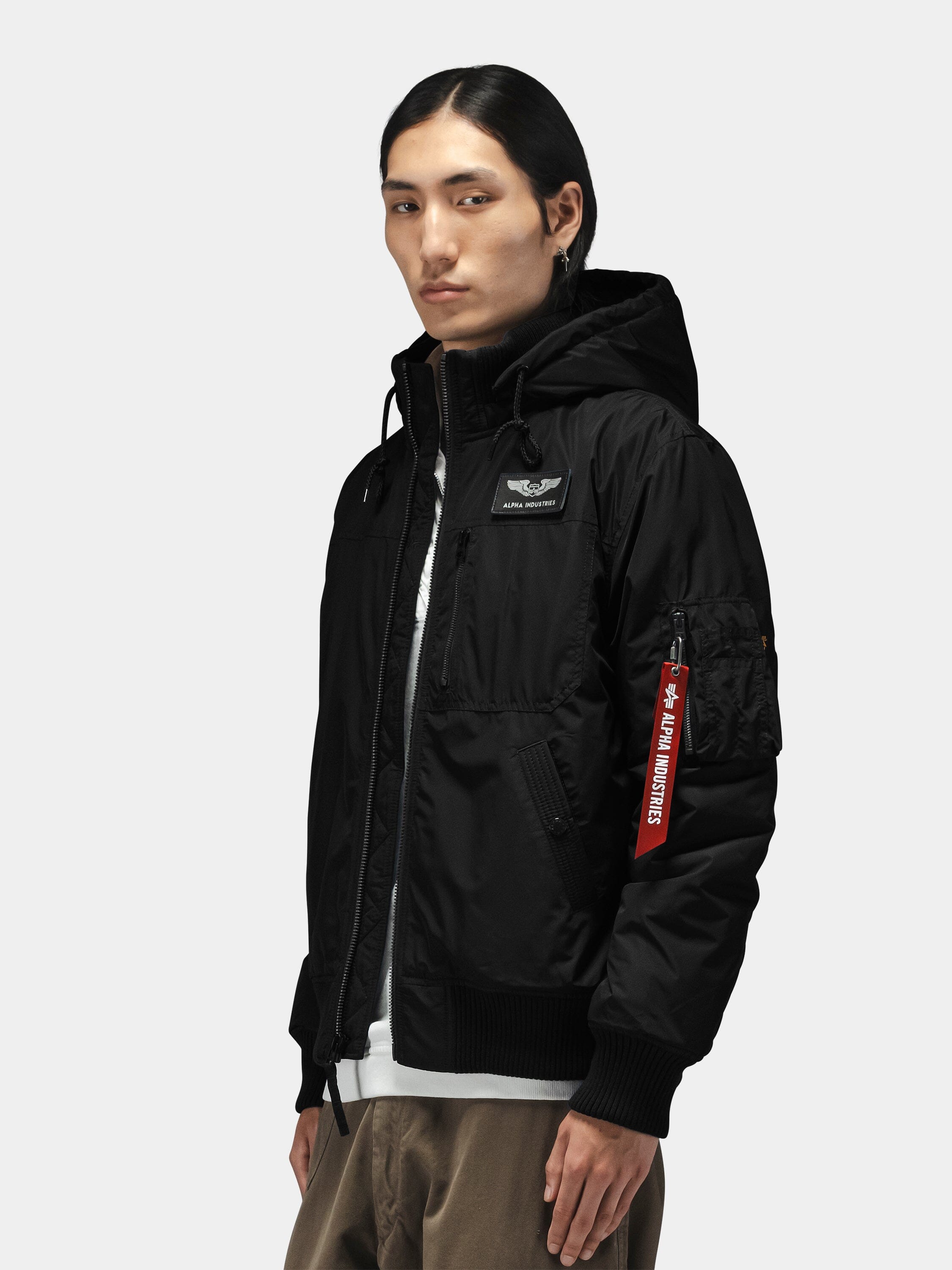 Alpha Industries Hooded Rib Flight Jacket with utility sleeve pocket, red tag, ribbed cuffs, layered over white shirt and khaki pants.