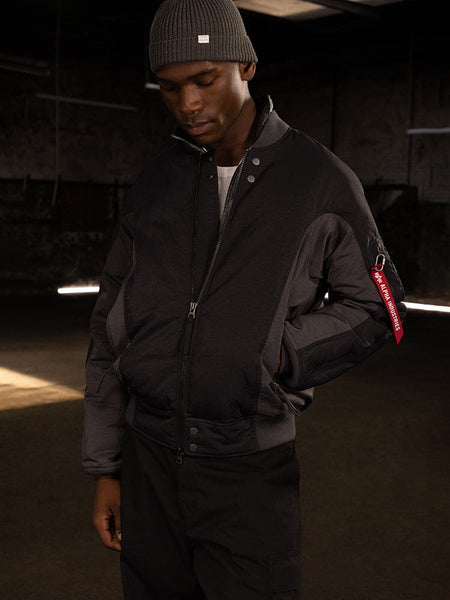 A person wearing an Alpha Industries Insulated MA-1 Bomber Jacket and a beanie stands, looking downward.