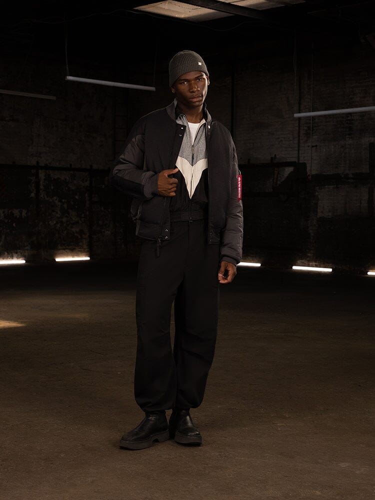 A person stands wearing the dark Insulated MA-1 Bomber Jacket by Alpha Industries, along with pants, a beanie, and boots.