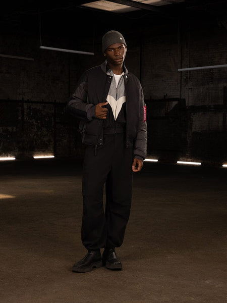 A person stands wearing the dark Insulated MA-1 Bomber Jacket by Alpha Industries, along with pants, a beanie, and boots.