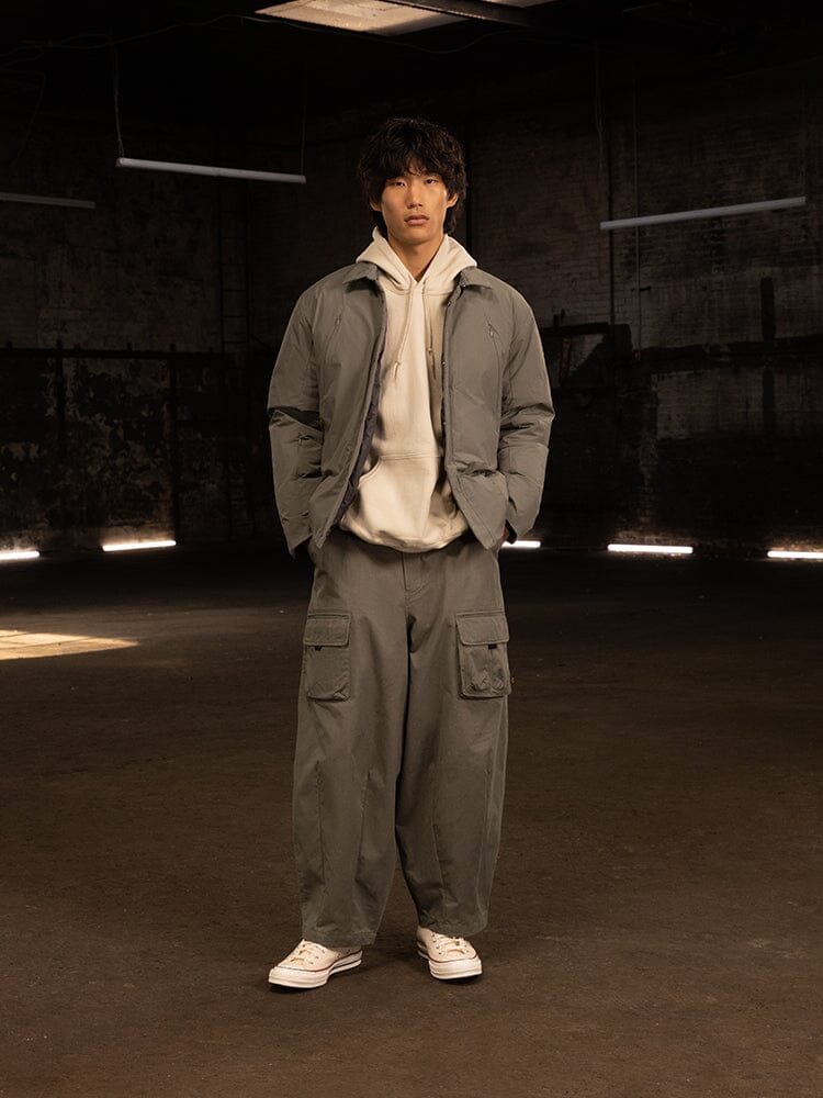 A person stands indoors wearing the water-resistant Insulated Shirt Jacket by Alpha Industries, paired with gray cargo pants, a white hoodie, and white sneakers. The streetwear ensemble is enhanced by industrial-style lighting.