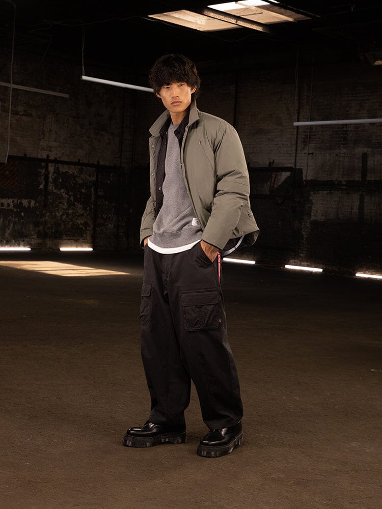 A person stands, sporting a streetwear look featuring the Insulated Shirt Jacket in gray by Alpha Industries, complemented by black cargo pants and matching black shoes.