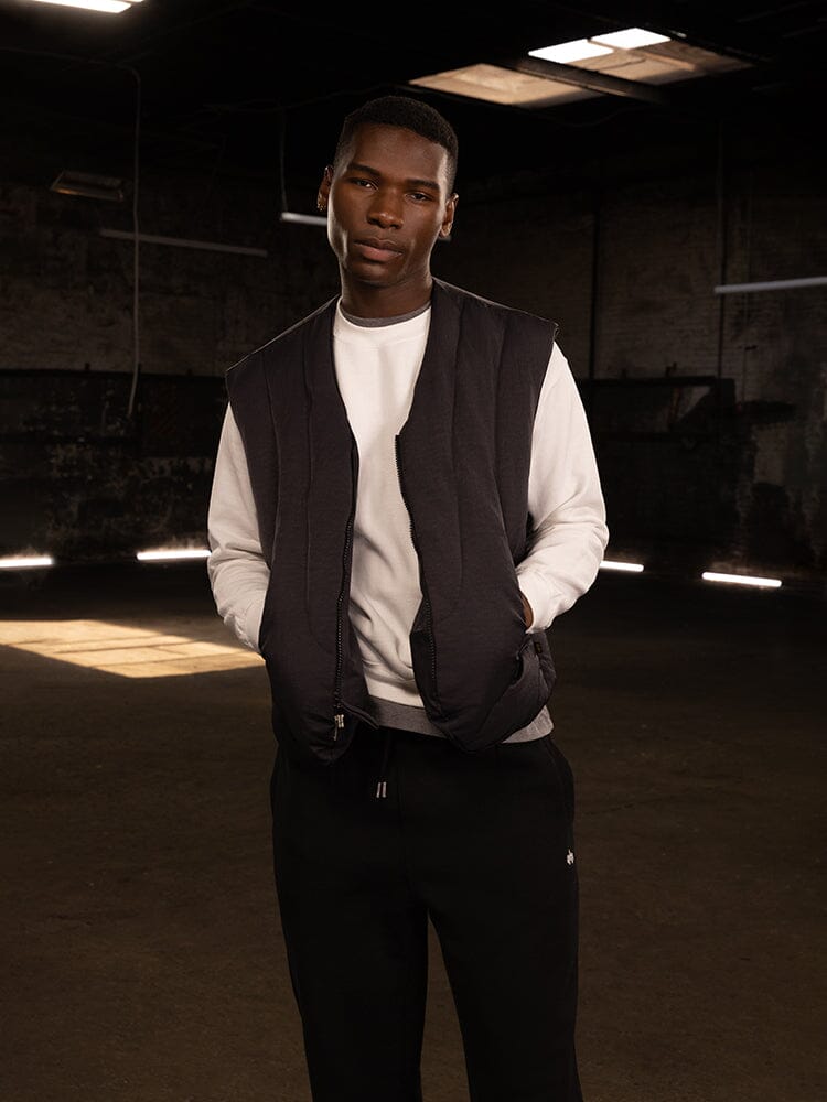 A person stands confidently with hands in the pockets of the Alpha Industries Insulated Vest, crafted from water-resistant nylon. Underneath is a down-filled layer made of responsible down fill, complemented by a crisp white shirt and sleek black pants.