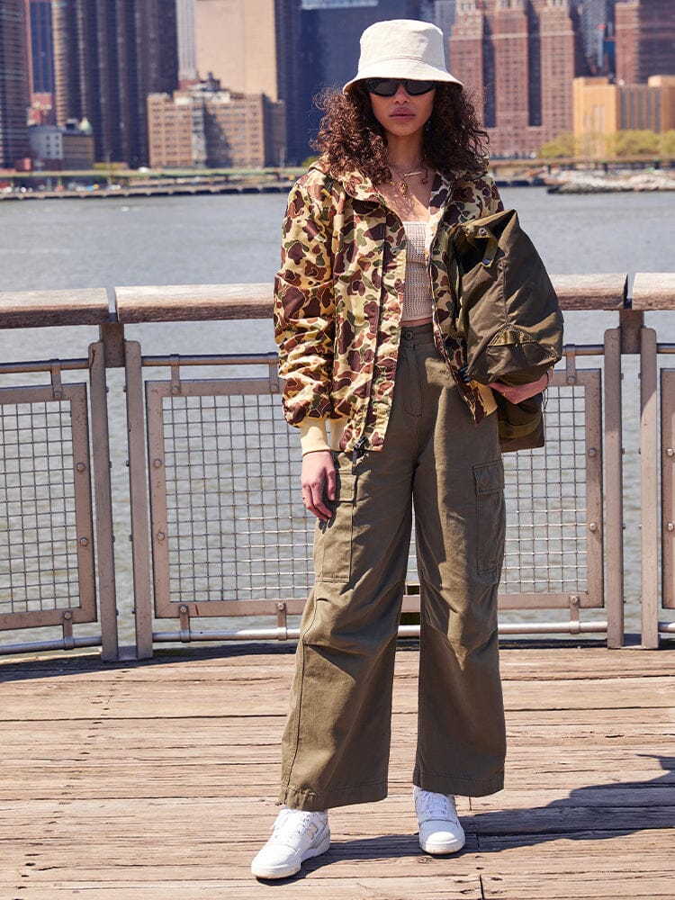 A person wearing a camo jacket, cargo pants, bucket hat, and sunglasses stands on a boardwalk. The ensemble is ideal for those who value style and functionality, with the option to swap to an Alpha Industries L-2B Hooded Gen II Bomber Jacket if the weather becomes unfavorable.