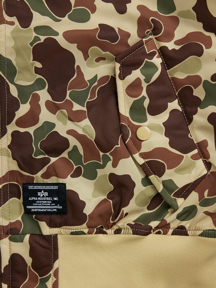 Close-up of the L-2B Hooded Gen II Bomber Jacket in camouflage nylon, showcasing a visible pocket and button. The black label displays "Alpha Industries, Inc." in white text.