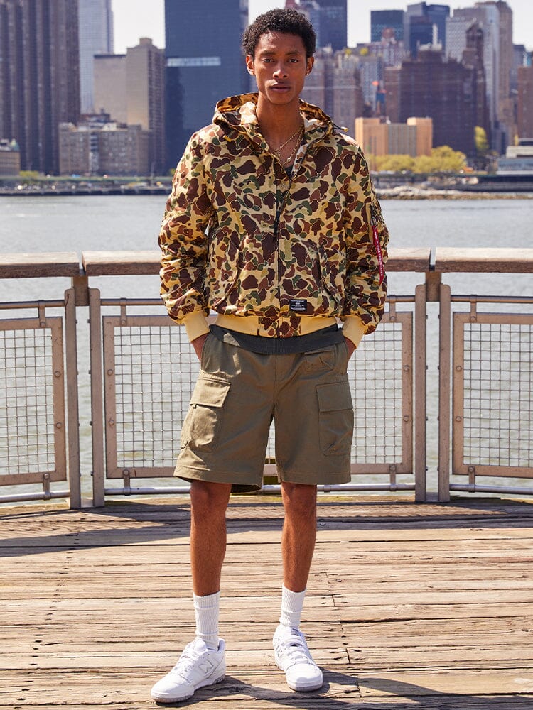 A person wearing an Alpha Industries L-2B Hooded Gen II Bomber Jacket, cargo shorts, and white sneakers stands on a wooden deck by the water.
