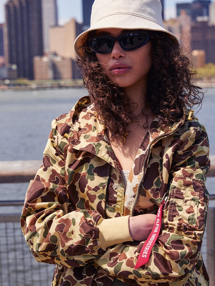 A person wearing an Alpha Industries L-2B Hooded Gen II Bomber Jacket, sunglasses, and a beige bucket hat stands outdoors.