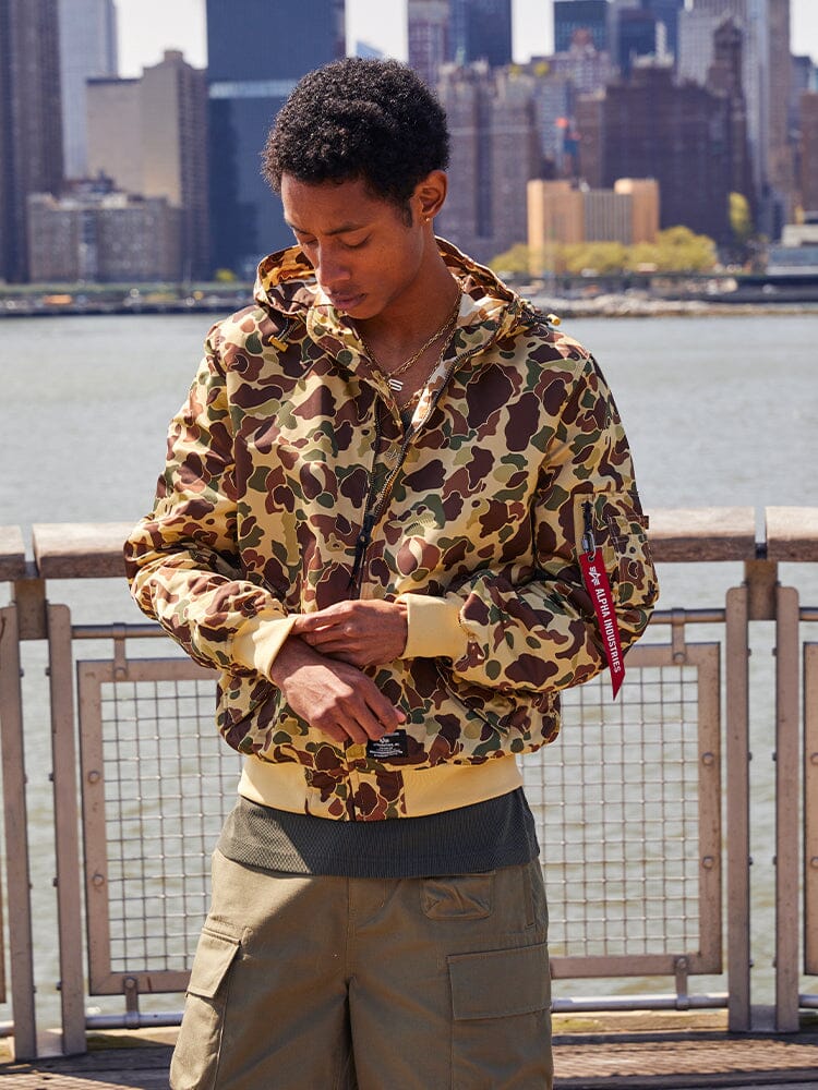 A person wearing an Alpha Industries L-2B Hooded Gen II Bomber Jacket and green pants stands on a waterfront.