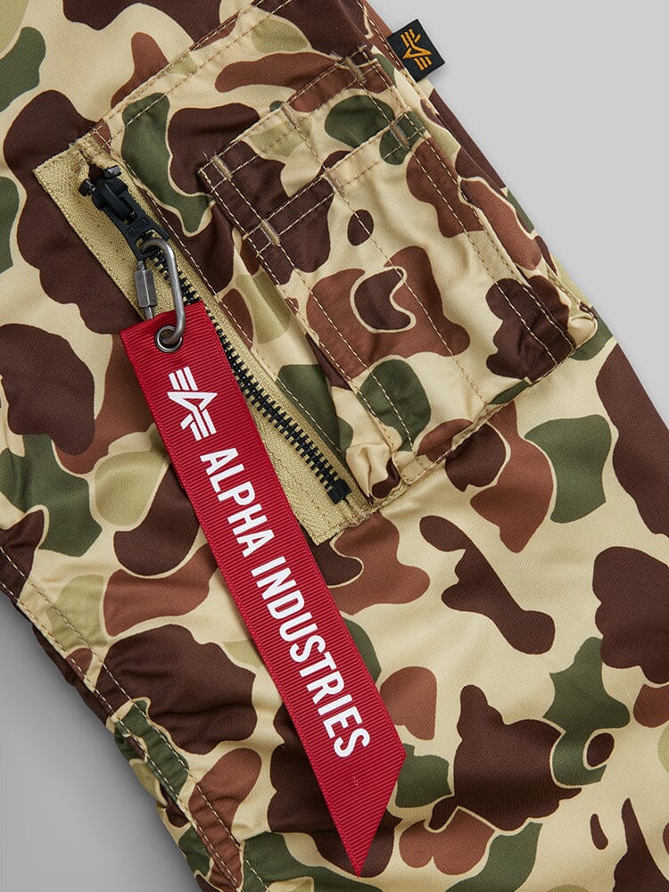 Camouflage cargo pants with a zipper pocket and a red Alpha Industries tag, expertly designed to pair flawlessly with the Alpha Industries L-2B Hooded Gen II Bomber Jacket.