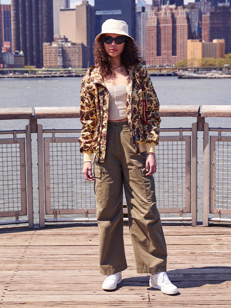 A person standing on a boardwalk is dressed in a camo jacket, beige tank top, khaki cargo pants, white sneakers, sunglasses, and a bucket hat. Their look combines urban vibes with the functionality of the L-2B Hooded Gen II Bomber Jacket by Alpha Industries, Inc., known for its water-resistant practicality.