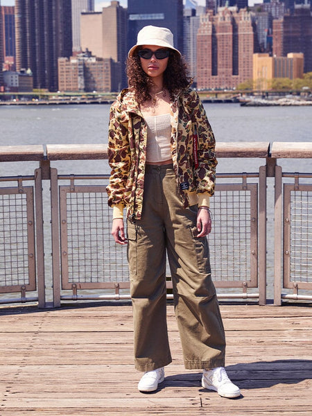 A person standing on a boardwalk is dressed in a camo jacket, beige tank top, khaki cargo pants, white sneakers, sunglasses, and a bucket hat. Their look combines urban vibes with the functionality of the L-2B Hooded Gen II Bomber Jacket by Alpha Industries, Inc., known for its water-resistant practicality.