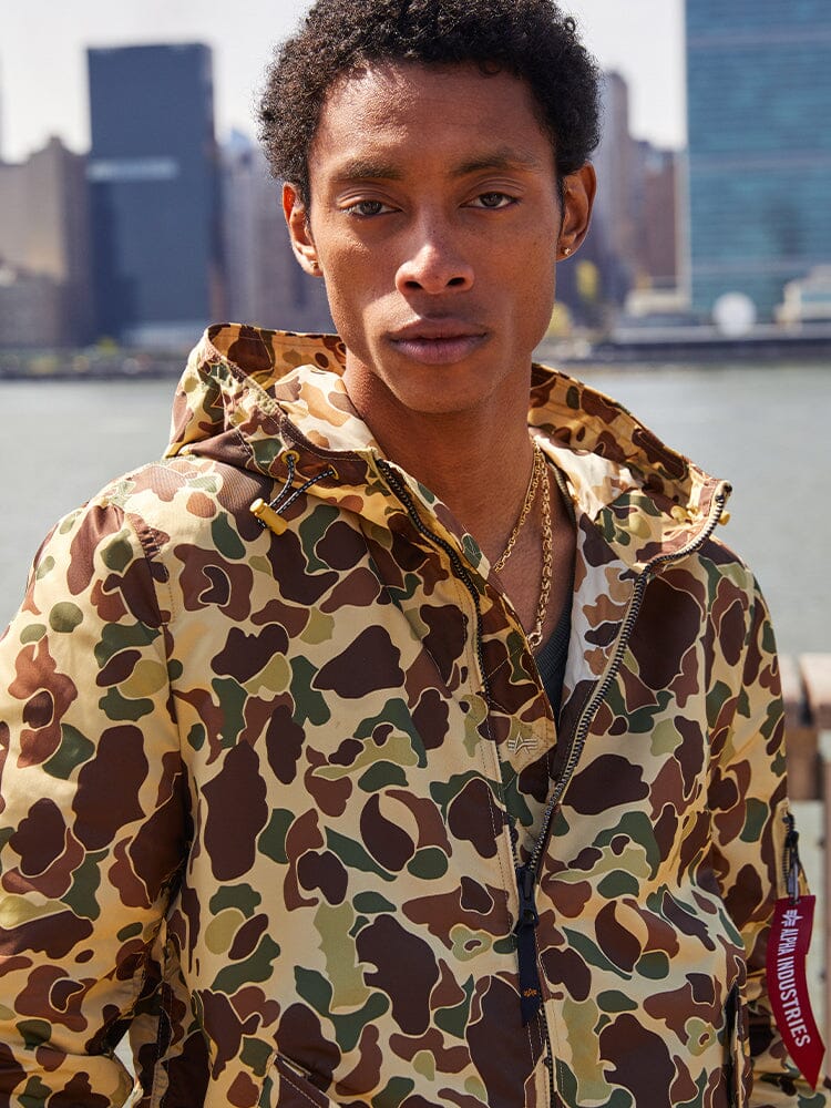 A person wearing an Alpha Industries L-2B Hooded Gen II Bomber Jacket stands outdoors.