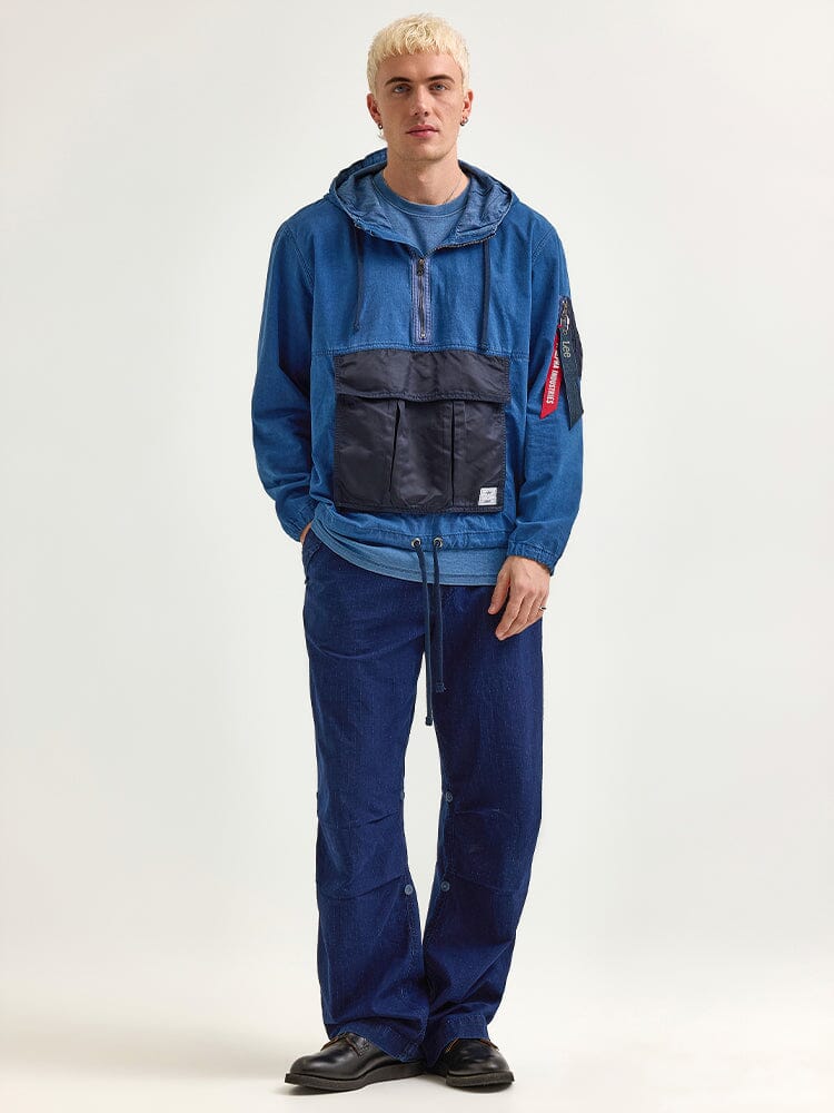 An individual dressed in the Alpha X Lee Anorak Jacket by Alpha Industries, with a blue denim jacket and pants that feature a black utility pocket and a red tag on the sleeve, poses.