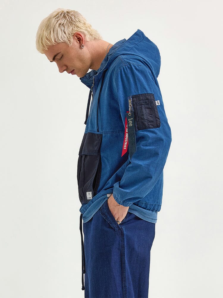 A person with blonde hair is wearing the Alpha X Lee Anorak Jacket from Alpha Industries, featuring a blue hood and utility pockets, along with jeans, while gazing downwards.