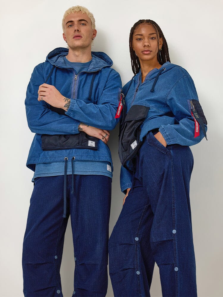 Two people stand side by side, each wearing Alpha X Lee Anorak Jackets in indigo, coordinated with matching denim pants and crossbody bags featuring utility pockets.