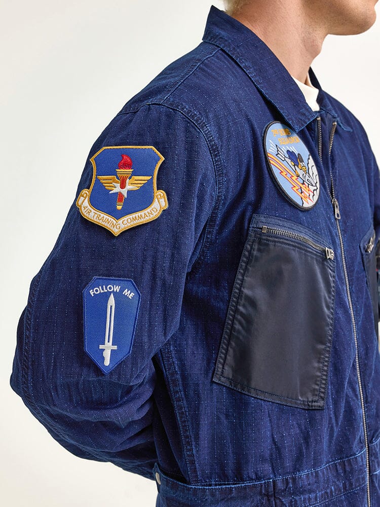 An individual wearing the Alpha x Lee Flight Suit, crafted from ripstop cotton with several patches such as Follow Me. This flight suit is a collaboration by Alpha Industries.