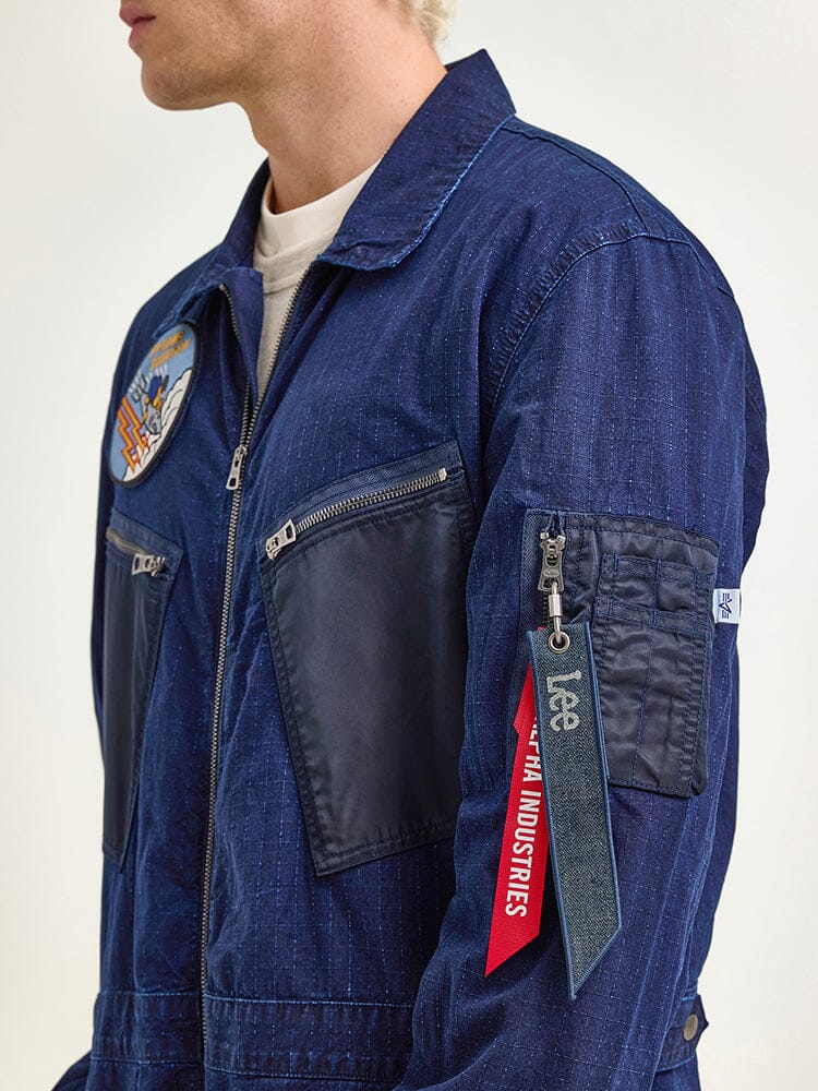 Dressed with confidence in the Alpha X Lee Flight Suit by Alpha Industries, this attire is made from durable ripstop cotton in blue, embellished with patches and showcasing a distinctive red Remove Before Flight tag on the sleeve. This blend of style and practicality represents performance fashion at its best.