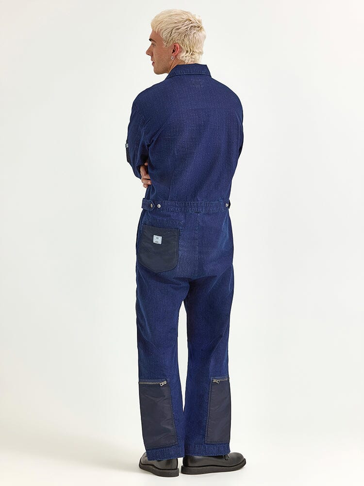 A person with short blonde hair, dressed in the Alpha X Lee Flight Suit from Alpha Industries, crafted from dark blue ripstop cotton, stands viewed from behind.