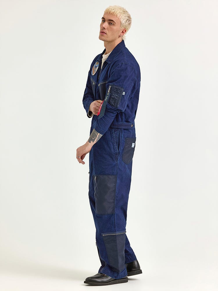 Wearing the Alpha X Lee Flight Suit by Alpha Industries, a person in a blue, patch-adorned design made from durable ripstop cotton stands sideways, epitomizing the essence of performance fashion.
