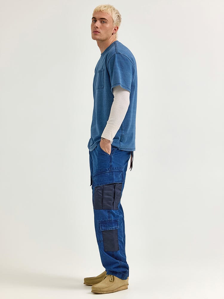 A person with short blonde hair stands in profile, wearing an Alpha X Lee Looser Fit Indigo Dyed Tee by Alpha Industries over a white long-sleeve, blue cargo pants with reinforced pockets, and brown shoes.