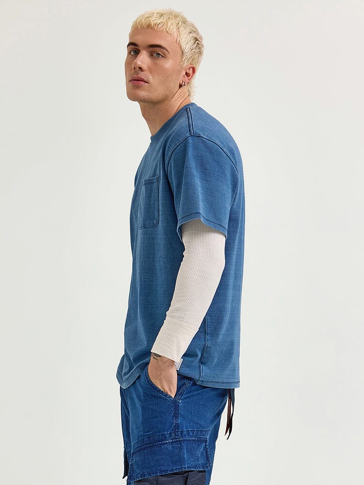 A person with short blond hair is wearing an Alpha Industries / Fw24-Lee Looser Fit Indigo Dyed Tee over a long-sleeve white shirt and indigo-dyed blue shorts, standing with one hand in a reinforced pocket.