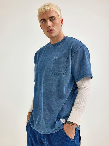 Dressed in an Indigo-Dyed Alpha X Lee Looser Fit Tee by Alpha Industries, the person layers it over a white long-sleeve shirt and has their hands tucked into reinforced pockets.