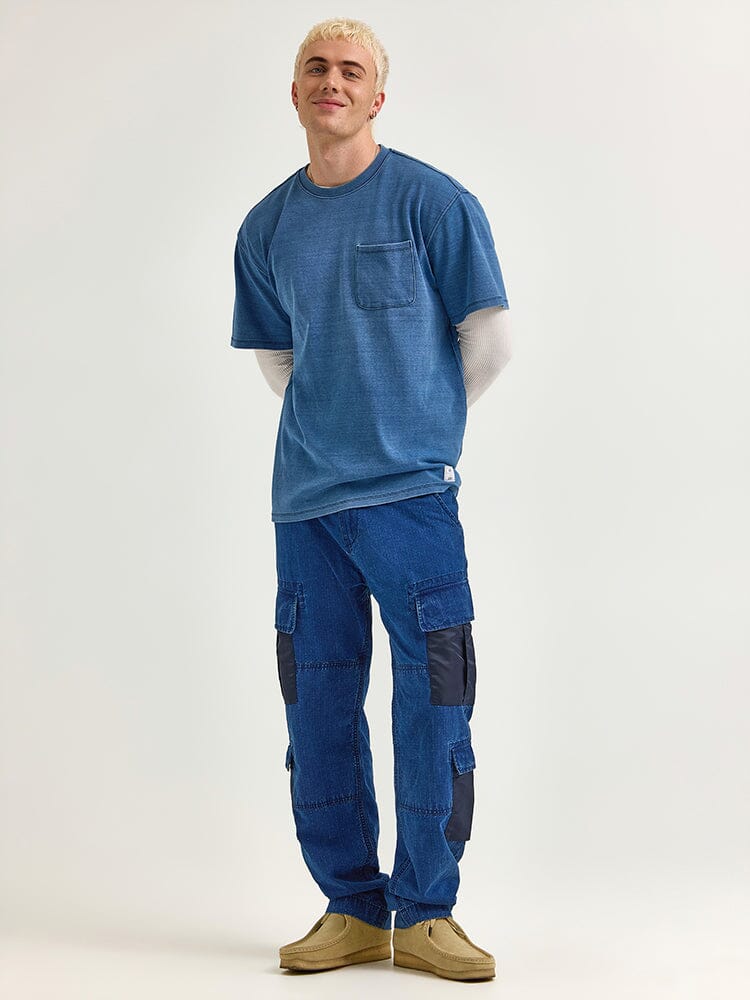 A person stands wearing the Alpha x Lee Looser Fit Indigo Dyed Tee by Alpha Industries over a long-sleeve shirt, complemented by blue cargo pants featuring reinforced pockets and patches, along with tan shoes.